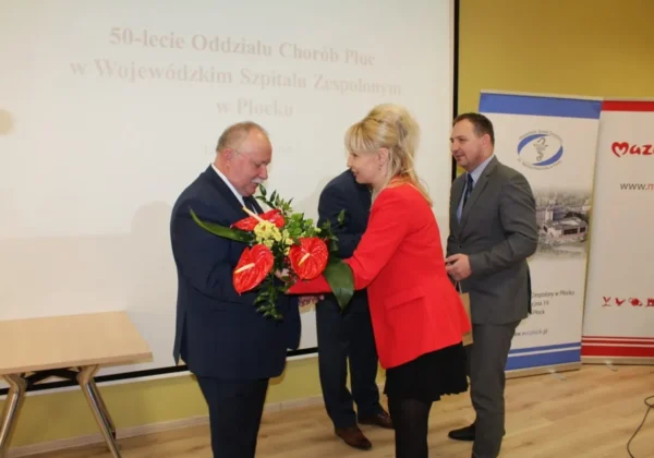 Mężczyzna w garniturze odbiera bukiet od kobiety w czerwonej marynarce podczas uroczystości 50-lecia Oddziału Chorób Płucnych w Szpitalu Wojewódzkim, na tle projekcji i bannerów.