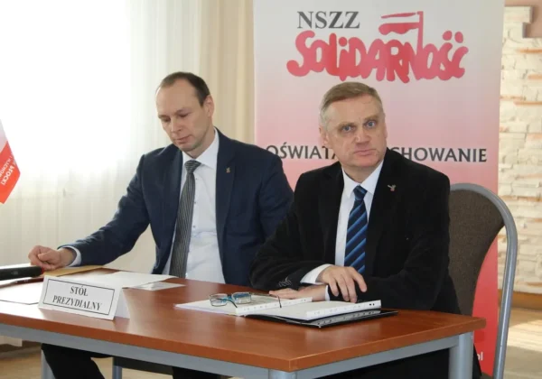 Dwóch mężczyzn w garniturach siedzi przy stole prezydialnym podczas sprawozdawczego zebrania Delegatów Sekcji Oświaty i Wychowania NSZZ Solidarność Regionu Płock, na tle banera z logo Solidarności.