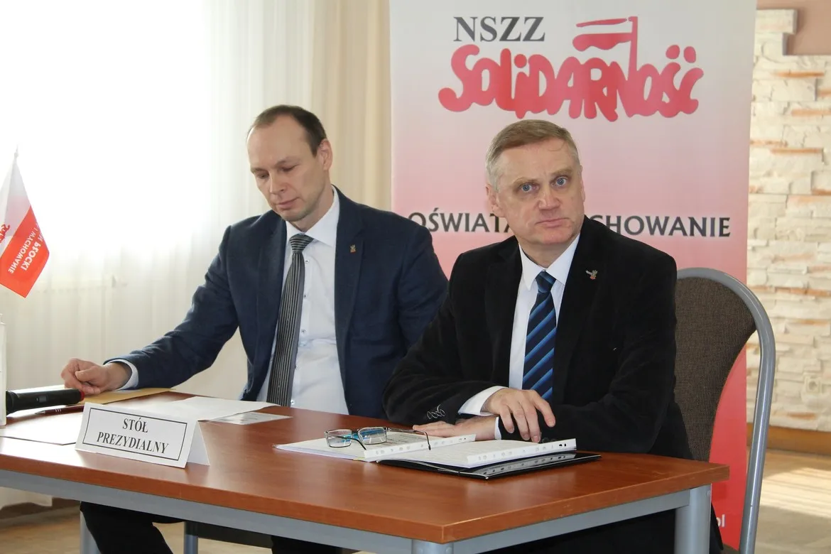 Dwóch mężczyzn w garniturach siedzi przy stole prezydialnym podczas sprawozdawczego zebrania Delegatów Sekcji Oświaty i Wychowania NSZZ Solidarność Regionu Płock, na tle banera z logo Solidarności.