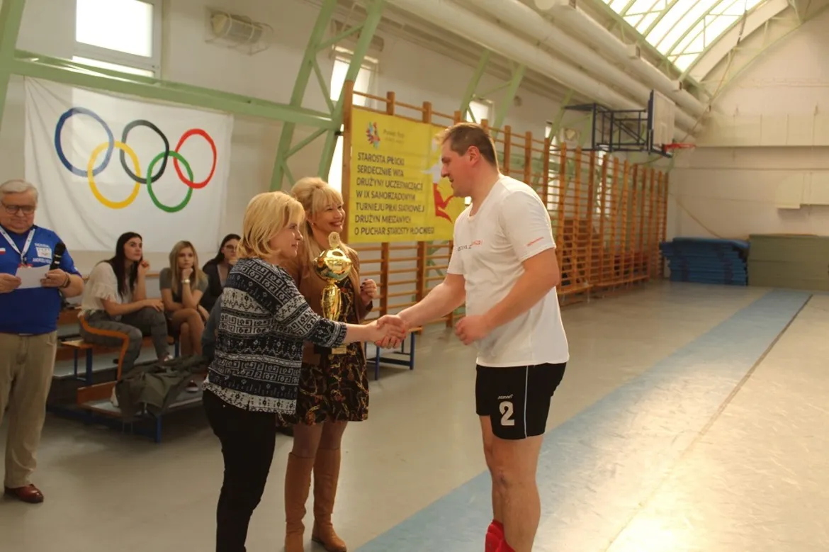 Podczas IX Turnieju Piłki Siatkowej Drużyn Mieszanych o Puchar Starosty Płockiego Mariusza Bieńka na sali gimnastycznej mężczyzna w białej koszulce odbiera puchar od kobiety, a w tle widać banner z olimpijskimi kręgami.