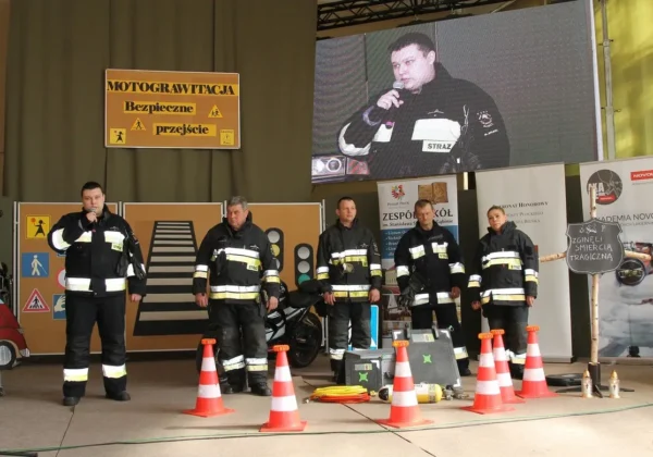 Grupa strażaków w pełnych mundurach stoi na scenie podczas prezentacji Motograwitacja – bezpieczne przejście, z tyłu plakatu z znakami drogowymi i zestawem sprzętu ratowniczego oraz pomarańczowymi stożkami.