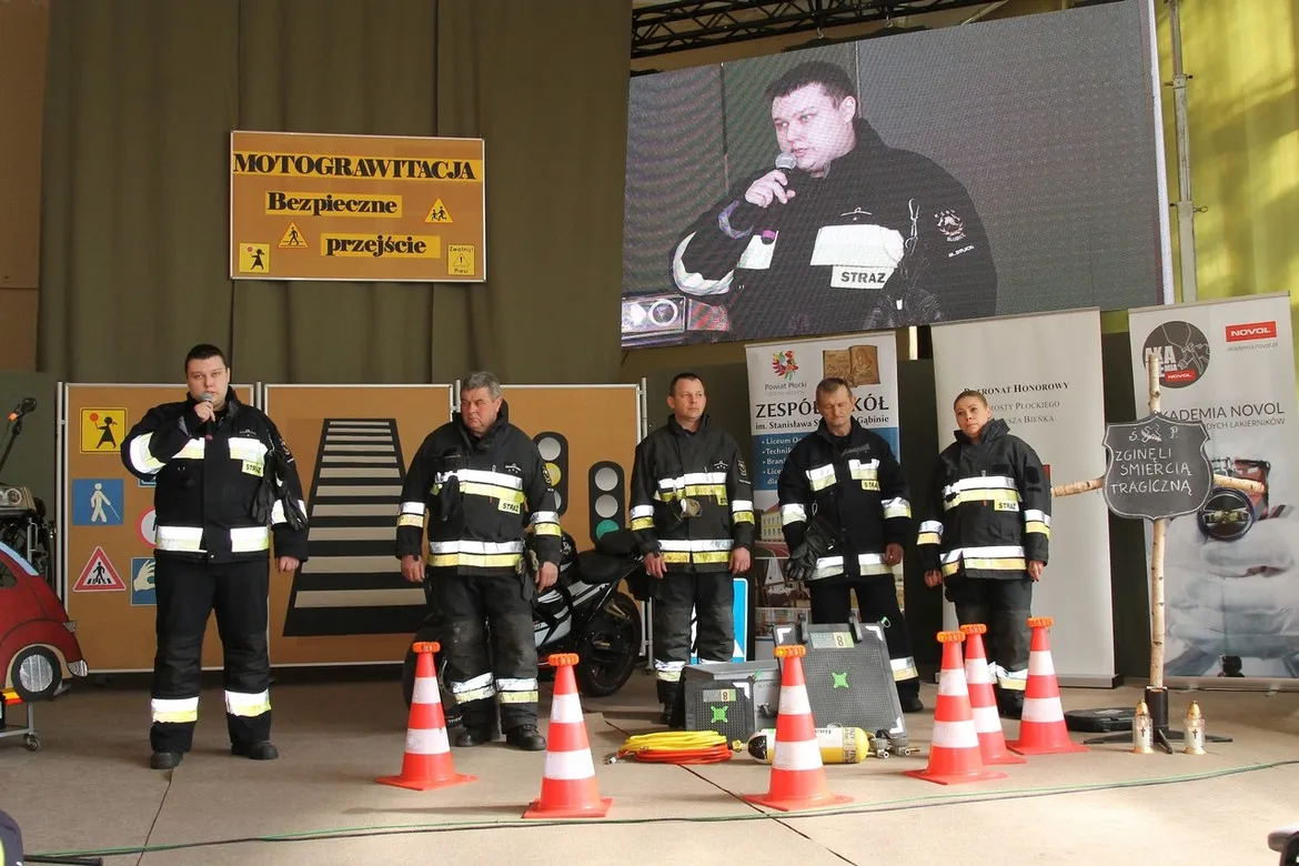 Grupa strażaków w pełnych mundurach stoi na scenie podczas prezentacji Motograwitacja – bezpieczne przejście, z tyłu plakatu z znakami drogowymi i zestawem sprzętu ratowniczego oraz pomarańczowymi stożkami.