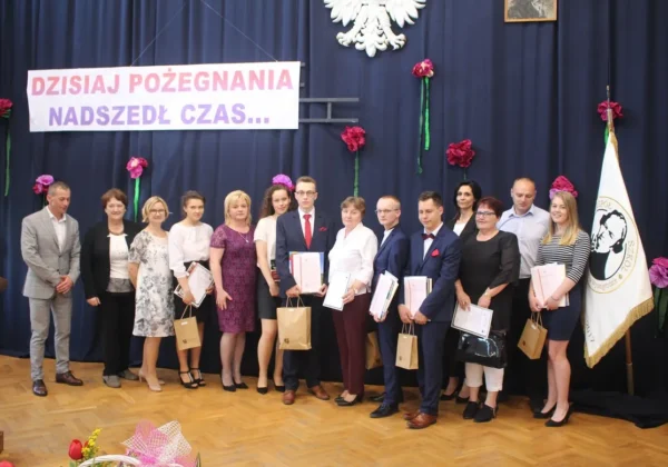 Grupa absolwentów w eleganckich strojach stoi na scenie podczas pożegnania, trzymając certyfikaty i torby prezentowe, w tle baner z napisem i flaga szkoły.