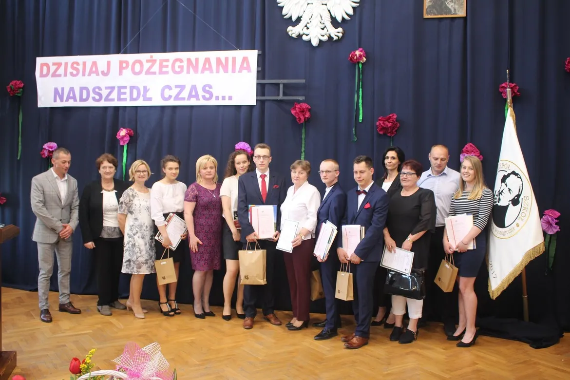 Grupa absolwentów w eleganckich strojach stoi na scenie podczas pożegnania, trzymając certyfikaty i torby prezentowe, w tle baner z napisem i flaga szkoły.