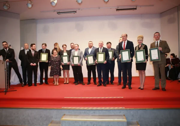 Grupa osób w formalnych strojach stoi na scenie podczas gali, każdy trzyma certyfikat w zielonej ramce.