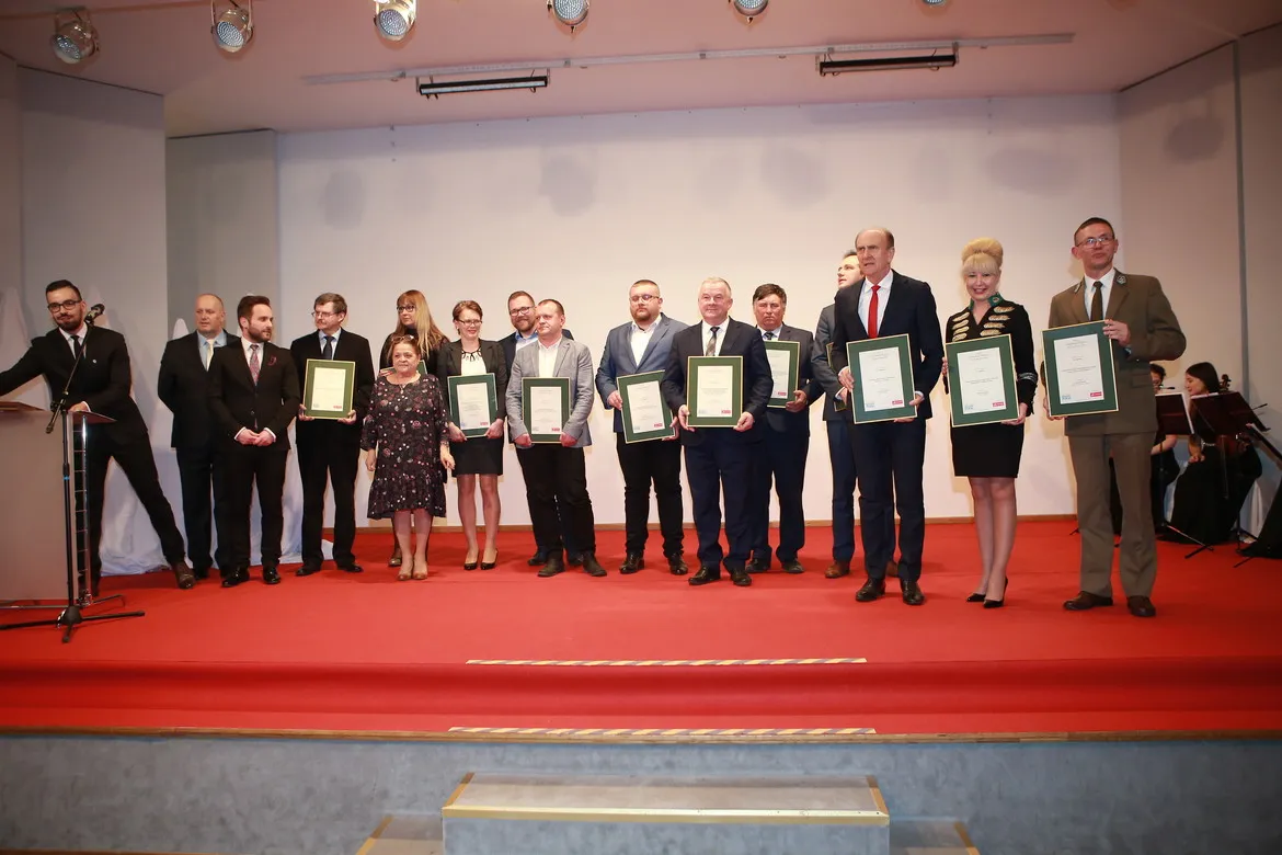 Grupa osób w formalnych strojach stoi na scenie podczas gali, każdy trzyma certyfikat w zielonej ramce.