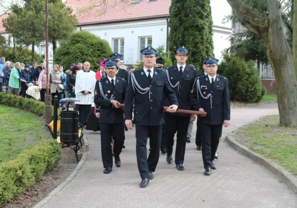 Czterech mężczyzn w mundurach niesie drewnianą belkę podczas Drogi Krzyżowej w DPS w Goślicach, za nimi podążają zgromadzeni uczestnicy.