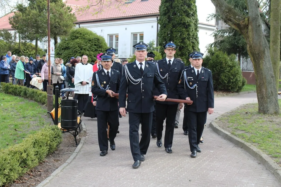 Czterech mężczyzn w mundurach niesie drewnianą belkę podczas Drogi Krzyżowej w DPS w Goślicach, za nimi podążają zgromadzeni uczestnicy.