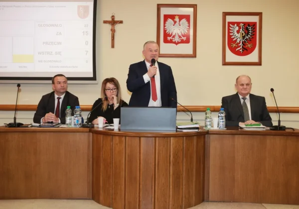 Podczas VII sesji Rady Powiatu cztery osoby siedzą przy drewnianym, półokrągłym stole; jeden mężczyzna stoi i przemawia do mikrofonu, a na ścianie widać krzyż i dwa herby, natomiast na projektorze wyświetlane są wyniki głosowania.