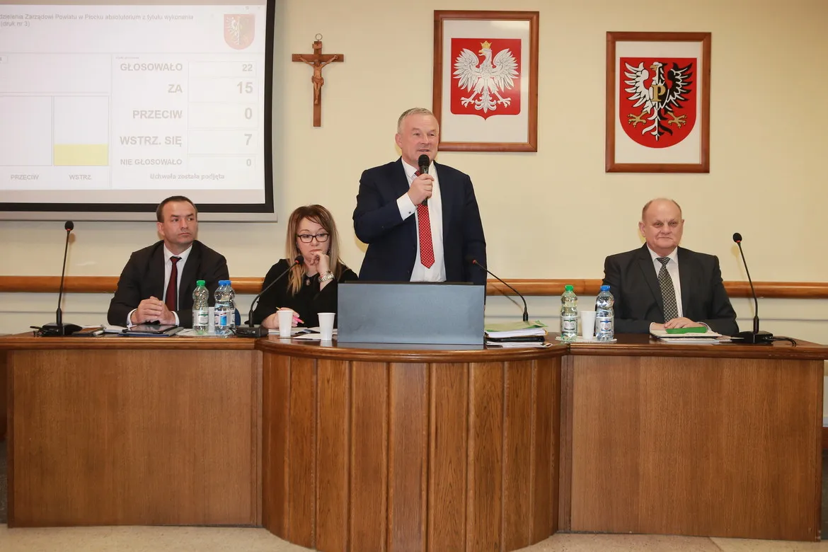 Podczas VII sesji Rady Powiatu cztery osoby siedzą przy drewnianym, półokrągłym stole; jeden mężczyzna stoi i przemawia do mikrofonu, a na ścianie widać krzyż i dwa herby, natomiast na projektorze wyświetlane są wyniki głosowania.