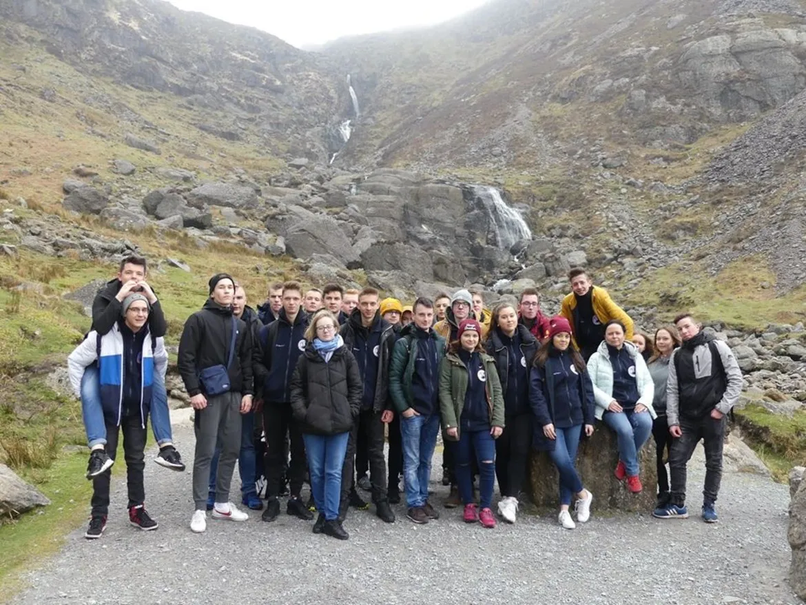 Grupa uczniów Zespołu Szkół im. Jana Śniadeckiego w Wyszogrodzie stoi na skalistej drodze w górach z wodospadem w tle podczas wyjazdu na praktyki zawodowe.