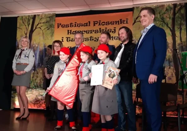 Na scenie podczas Festiwalu Piosenki Harcerskiej i Turystycznej grupa dzieci w harcerskich mundurach z czerwonymi beretami prezentuje nagrody, a w tle widoczny jest baner.