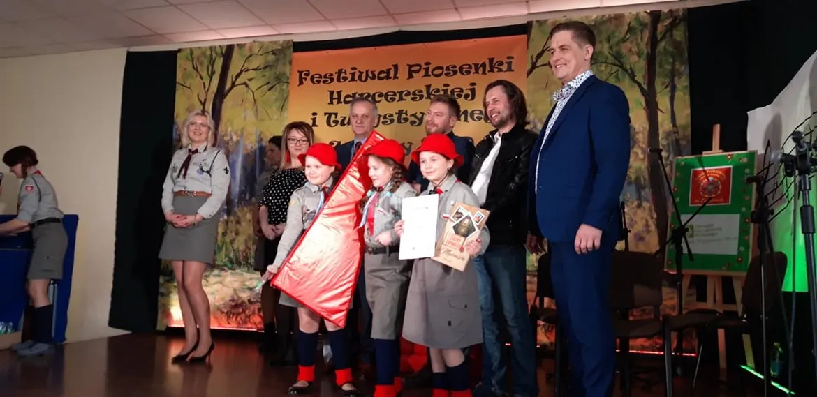Na scenie podczas Festiwalu Piosenki Harcerskiej i Turystycznej grupa dzieci w harcerskich mundurach z czerwonymi beretami prezentuje nagrody, a w tle widoczny jest baner.