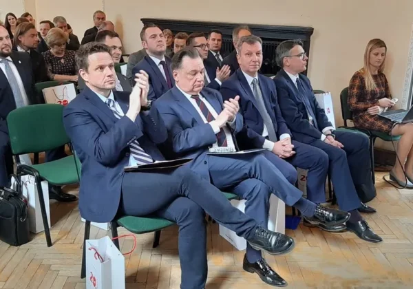 Grupa mężczyzn w garniturach siedzi w pierwszym rzędzie sali konferencyjnej podczas konferencji „Forum dla Mazowsza – Mazowsze w Europie po 2020 r.”, część z nich klaska, a po prawej stronie stoi osoba robiąca zdjęcie.