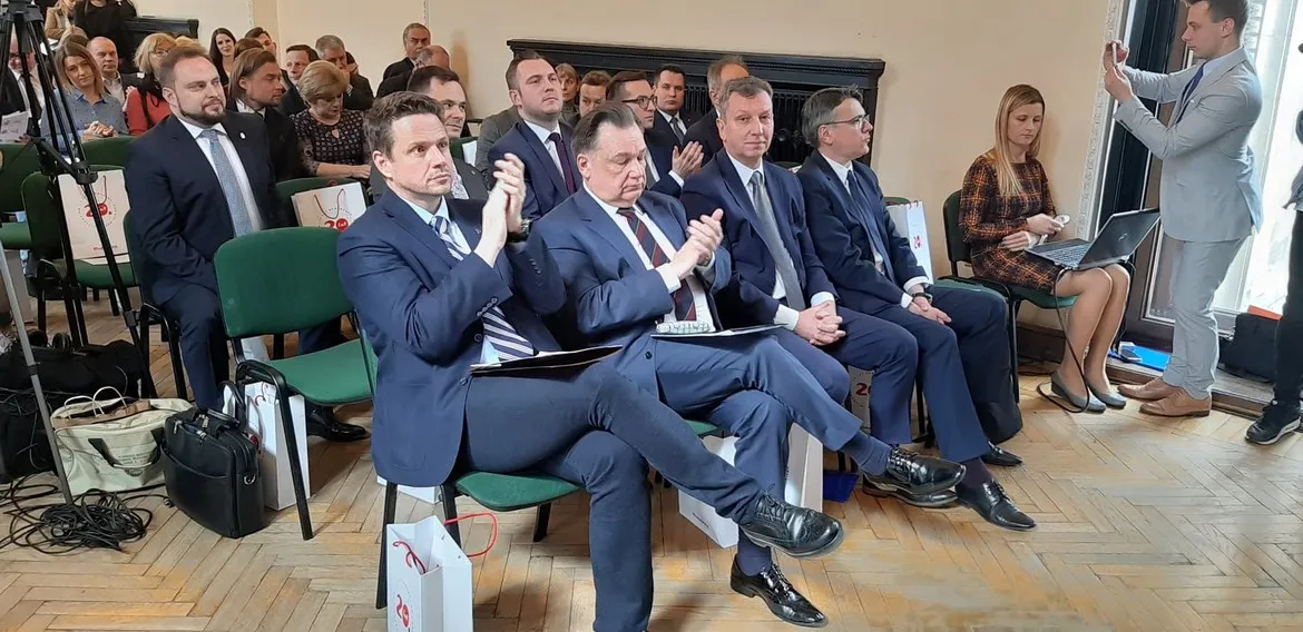 Grupa mężczyzn w garniturach siedzi w pierwszym rzędzie sali konferencyjnej podczas konferencji „Forum dla Mazowsza – Mazowsze w Europie po 2020 r.”, część z nich klaska, a po prawej stronie stoi osoba robiąca zdjęcie.