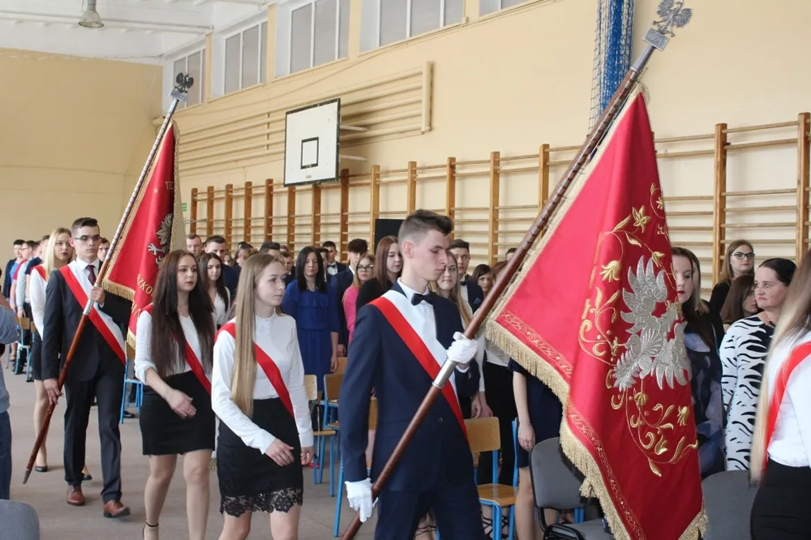Maturzyści z Gąbina maszerują z flagami podczas uroczystości zakończenia roku szkolnego w szkolnej hali gimnastycznej.