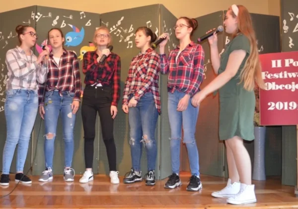 Sześć dziewcząt stoi na scenie podczas II Powiatowego Festiwalu Piosenki Obcojęzycznej, śpiewając do mikrofonów.