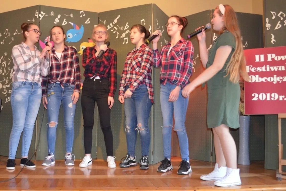 Sześć dziewcząt stoi na scenie podczas II Powiatowego Festiwalu Piosenki Obcojęzycznej, śpiewając do mikrofonów.