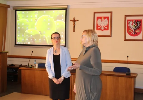 Dwie kobiety stojące w sali konferencyjnej płockiego starostwa rozmawiają podczas spotkania „Odczarować adopcję”, na tle projektora, mikrofonów oraz krzyża i herbów na ścianie.