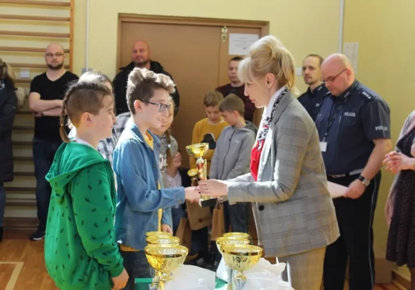 Na zdjęciu młodzi uczestnicy powiatowych eliminacji Turnieju BRD odbierają nagrody od kobiety w garniturze, stojąc przy stole z pucharami podczas ceremonii w sali gimnastycznej.