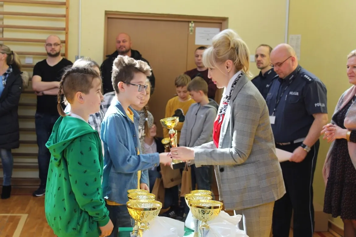 Na zdjęciu młodzi uczestnicy powiatowych eliminacji Turnieju BRD odbierają nagrody od kobiety w garniturze, stojąc przy stole z pucharami podczas ceremonii w sali gimnastycznej.
