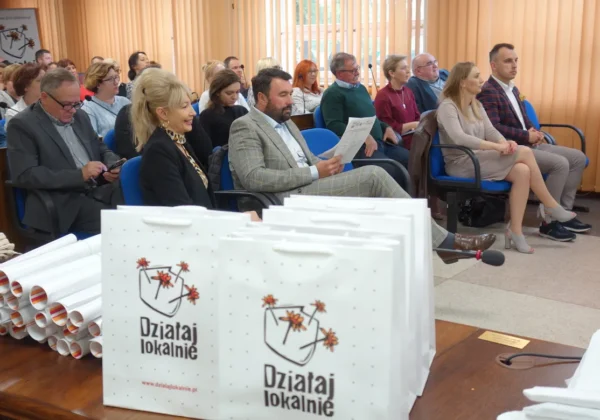 Grupa uczestników siedzi w sali konferencyjnej podczas ogłaszania wyników konkursu grantowego „Działaj Lokalnie”; na pierwszym planie widoczne są białe torby z logo „Działaj lokalnie” oraz zrolowane plakaty.