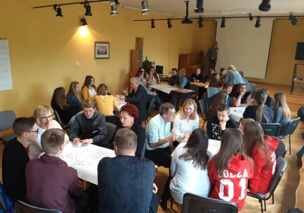 Uczestnicy zajęć integracyjnych Szkoły Dialogu w Zespole Szkół w Gąbinie siedzą w grupach przy stolikach, dyskutując i notując na arkuszach papieru.