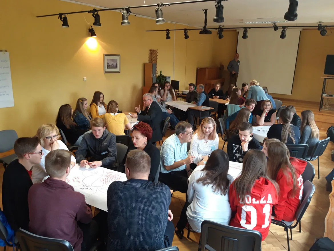 Uczestnicy zajęć integracyjnych Szkoły Dialogu w Zespole Szkół w Gąbinie siedzą w grupach przy stolikach, dyskutując i notując na arkuszach papieru.