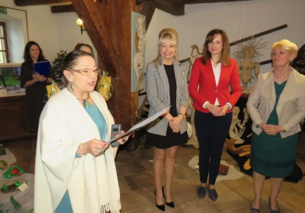 Grupa osób na wernisażu wystawy plenerowej „Mazowiecka Wieś 2019” w Nowym Miszewie; kobieta ubrana w biały ponczo wygłasza przemówienie.