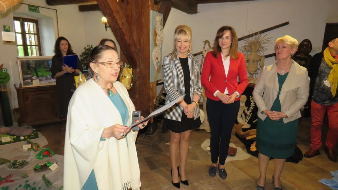 Grupa osób na wernisażu wystawy plenerowej „Mazowiecka Wieś 2019” w Nowym Miszewie; kobieta ubrana w biały ponczo wygłasza przemówienie.