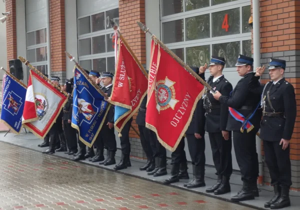Grupa strażaków PSP i OSP stoi na placu przed budynkiem, trzymając flagi podczas obchodów Dnia Strażaka w Płocku.
