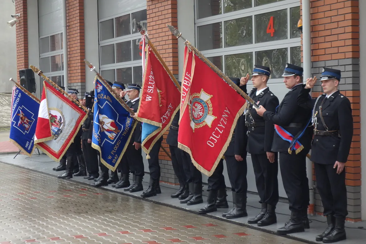 Grupa strażaków PSP i OSP stoi na placu przed budynkiem, trzymając flagi podczas obchodów Dnia Strażaka w Płocku.
