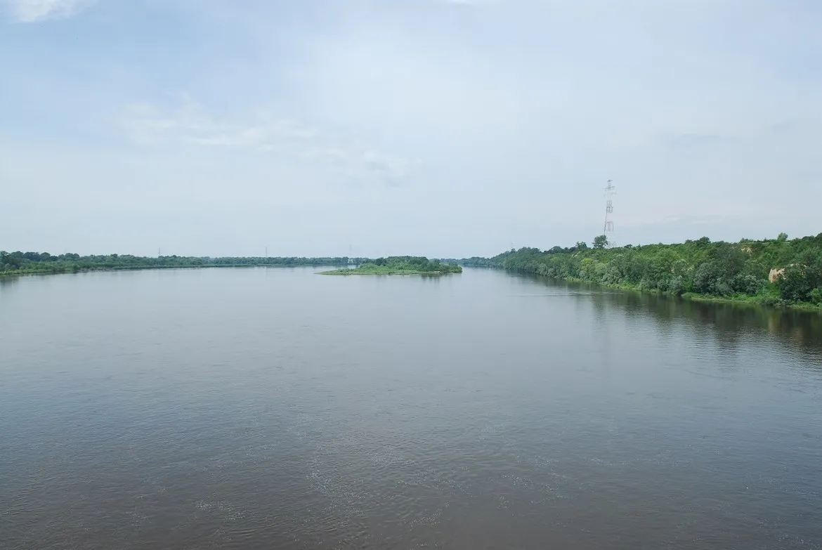 Szeroka rzeka z zielonymi brzegami i wyspą na środku pod jasnym niebem, nadciąga fala wezbraniowa w stronę Płocka.