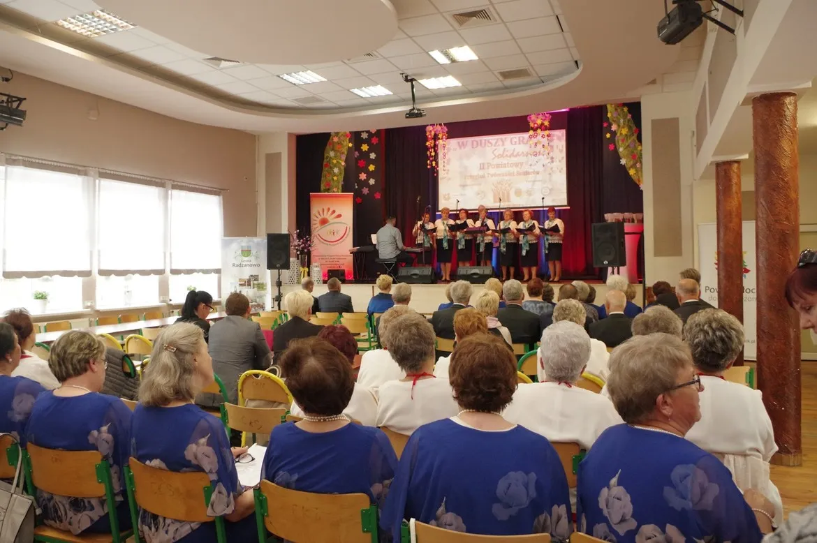 Na scenie w Radzanowie chór seniorów wykonuje występ podczas II Powiatowego Przeglądu Twórczości Seniorów, przed liczną widownią seniorów.