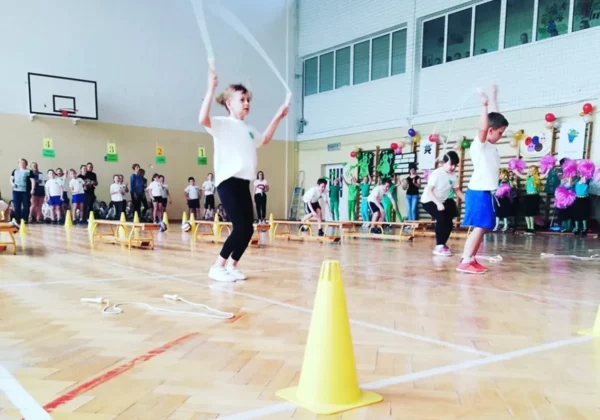 Dwoje dzieci skacze przez skakankę podczas turnieju w Nowym Miszewie w hali sportowej, przed widzami i kolorowymi dekoracjami.