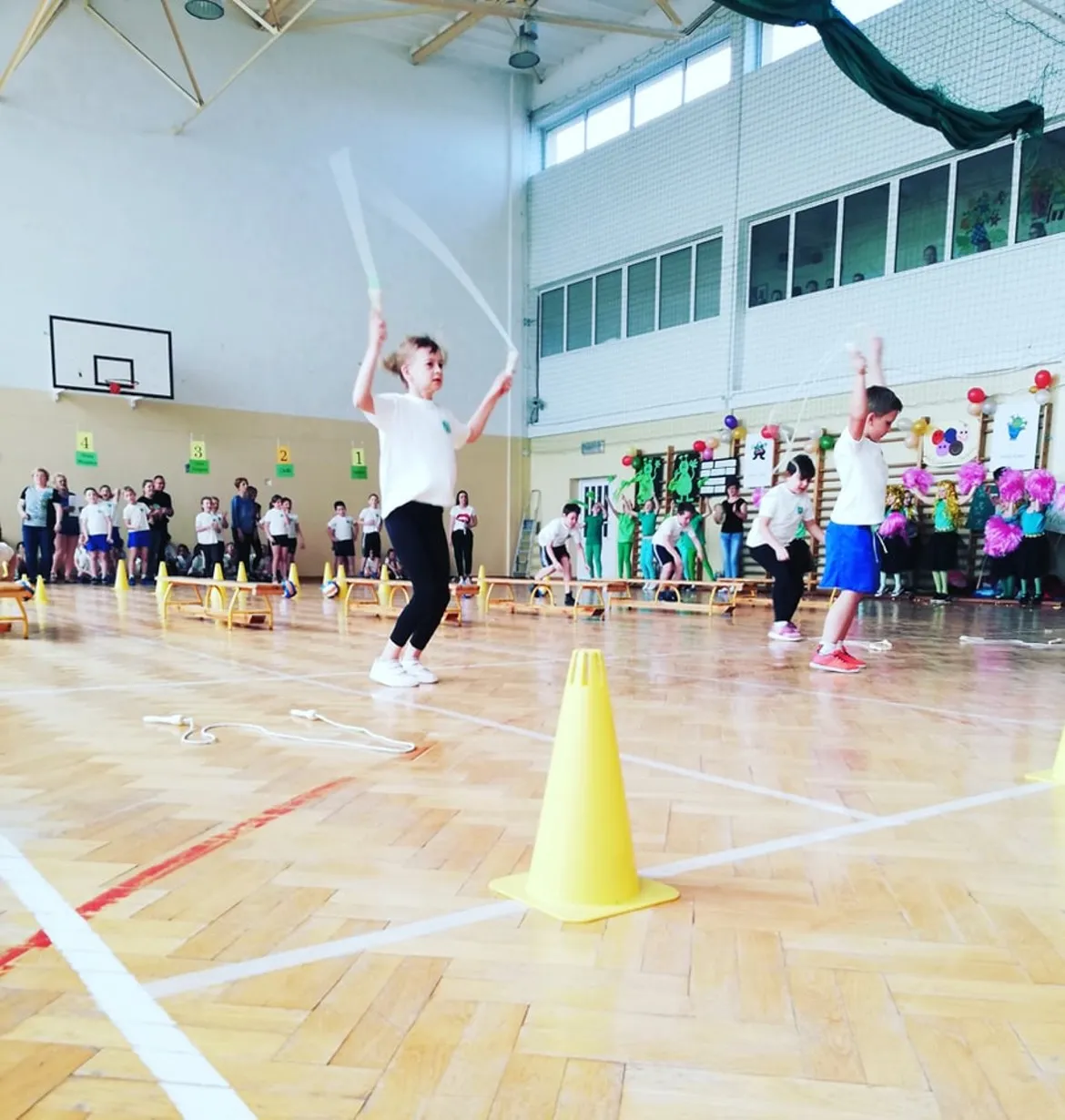 Dwoje dzieci skacze przez skakankę podczas turnieju w Nowym Miszewie w hali sportowej, przed widzami i kolorowymi dekoracjami.