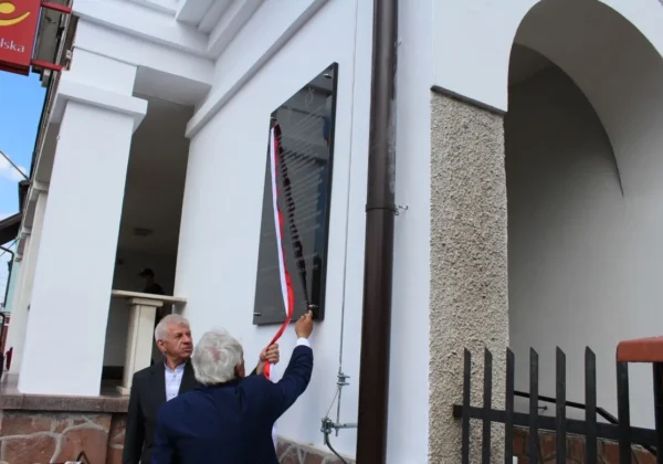 Dwóch starszych mężczyzn odsłania tablicę pamiątkową ku czci byłych mieszkańców Ziemi Wyszogrodzkiej, umieszczoną na białej elewacji budynku.