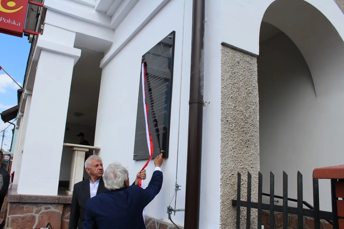 Dwóch starszych mężczyzn odsłania tablicę pamiątkową ku czci byłych mieszkańców Ziemi Wyszogrodzkiej, umieszczoną na białej elewacji budynku.