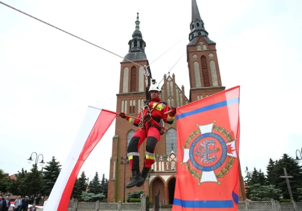 Strażak w czerwonym mundurze i hełmie zawieszony na linach podczas pokazu przed kościołem z dwiema wieżami, trzymający flagi.
