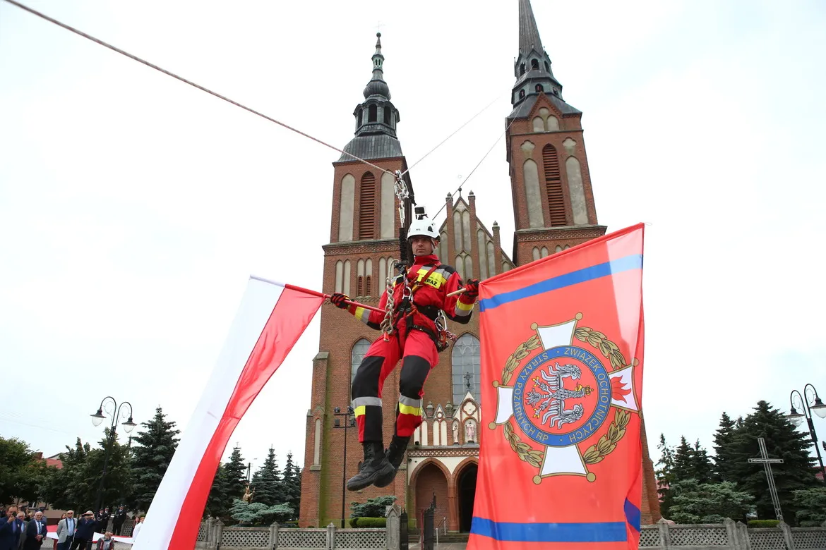 Strażak w czerwonym mundurze i hełmie zawieszony na linach podczas pokazu przed kościołem z dwiema wieżami, trzymający flagi.