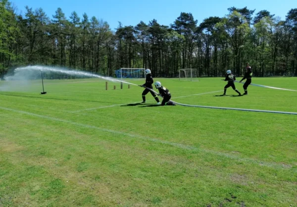Strażacy OSP w pełnym umundurowaniu gaszą wodę z węża podczas zawodów sportowo-pożarniczych na zielonym boisku.