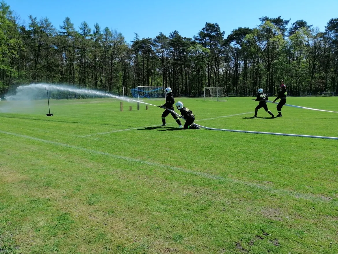 Strażacy OSP w pełnym umundurowaniu gaszą wodę z węża podczas zawodów sportowo-pożarniczych na zielonym boisku.