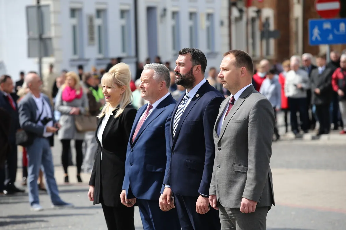 Czwórka uczestników uroczystości w formalnych garniturach stoi w rzędzie na ulicy podczas obchodów 228. rocznicy Konstytucji 3 Maja.