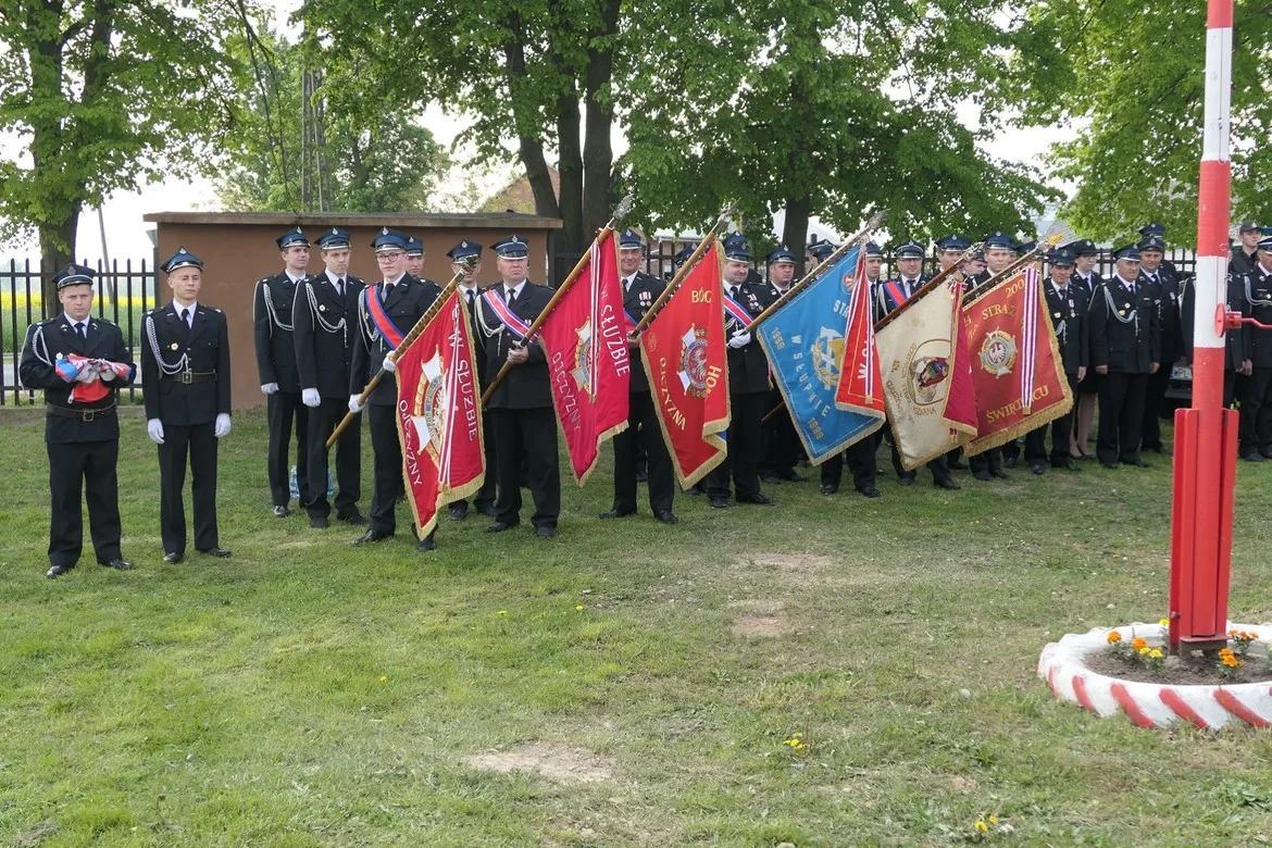 Grupa strażaków w mundurach służbowych stoi w szeregu i trzyma barwne sztandary podczas Dnia Strażaka w gminie Słupno.