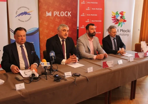 Czterech mężczyzn siedzi przy stole podczas konferencji prasowej w ramach Płockiego Forum Biznesu, z mikrofonami, notatkami i banerami z logo miasta Płock w tle.