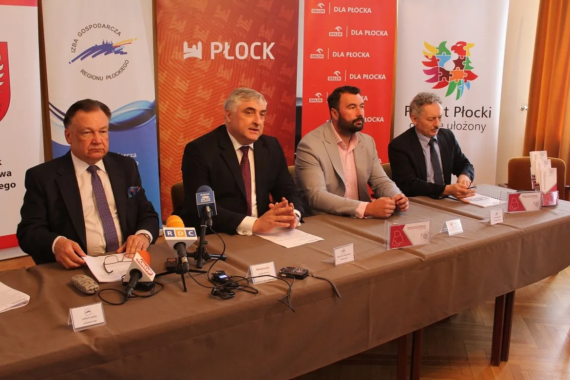 Czterech mężczyzn siedzi przy stole podczas konferencji prasowej w ramach Płockiego Forum Biznesu, z mikrofonami, notatkami i banerami z logo miasta Płock w tle.