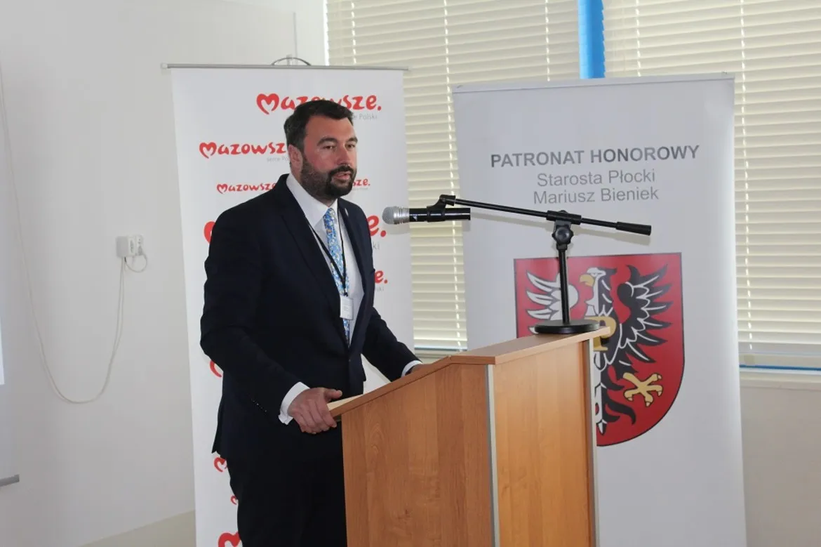 Mężczyzna w ciemnym garniturze przemawia z podiumu podczas Ogólnopolskiej Konferencji Naukowej „Pracownik nie praca, nie ucieknie? Współczesne tendencje na rynku pracy”, na tle banerów z napisem Mazowsze i herbem powiatu.