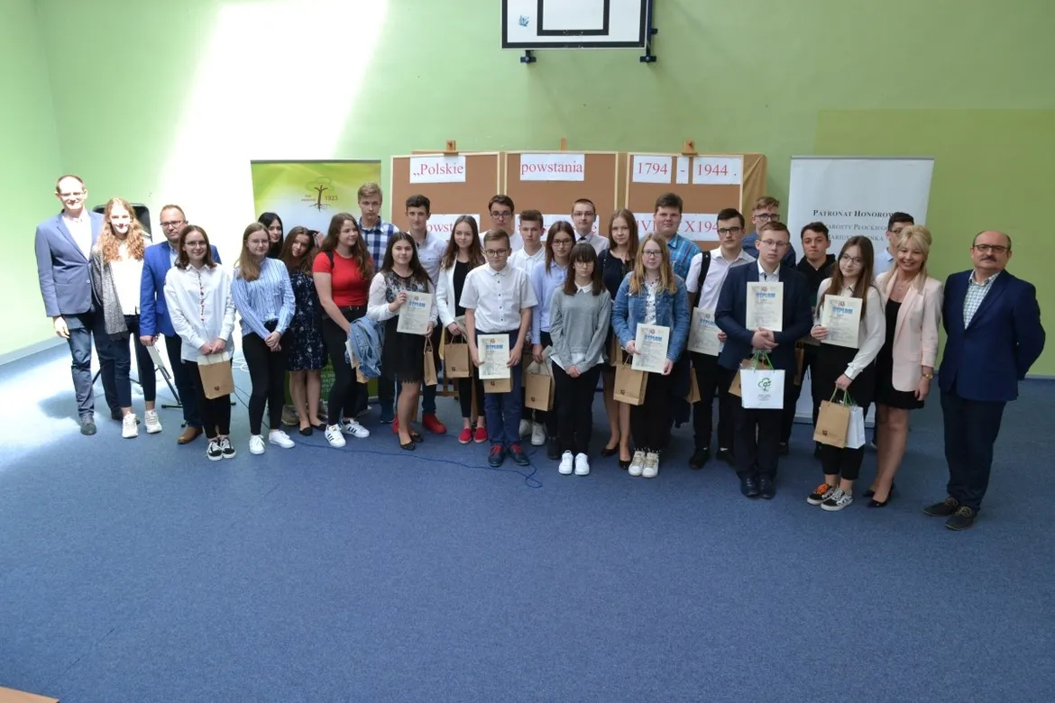 Grupa uczniów i nauczycieli stojąca razem w sali podczas wręczania nagród w konkursie historycznym „Polskie powstania 1794-1944”.