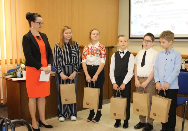 Grupa dzieci wraz z dorosłą kobietą stoi na sali podczas Dnia Rodzicielstwa Zastępczego, trzymając papierowe torby z upominkami.