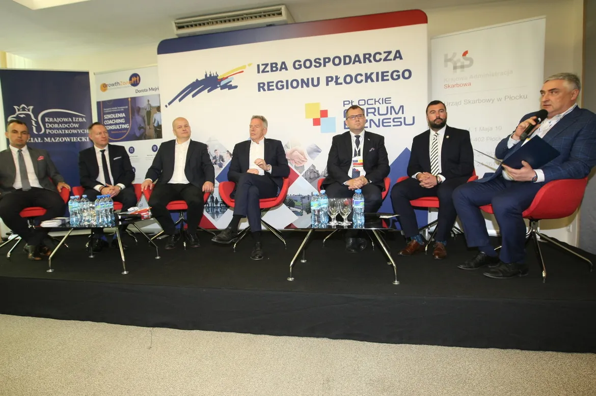 Siedmioosobowy panel dyskusyjny: mężczyźni w garniturach siedzą na czerwonych krzesłach na scenie z banerami Izby Gospodarczej Regionu Płockiego w tle.